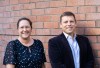 Rachel Perret and Rob Weinkove, smiling in front of a brick wall. Rachel (left) wears glasses and a dark spotted blouse; Rob (right) wears a blazer over an open-collared shirt.